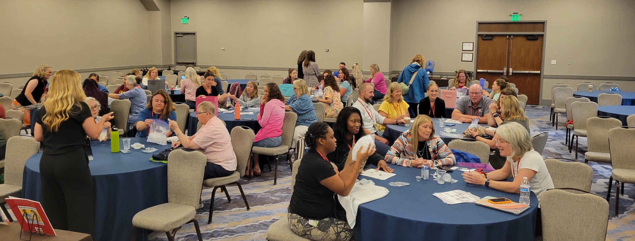 Large room with many round tables. People sitting at tables doing an activity.