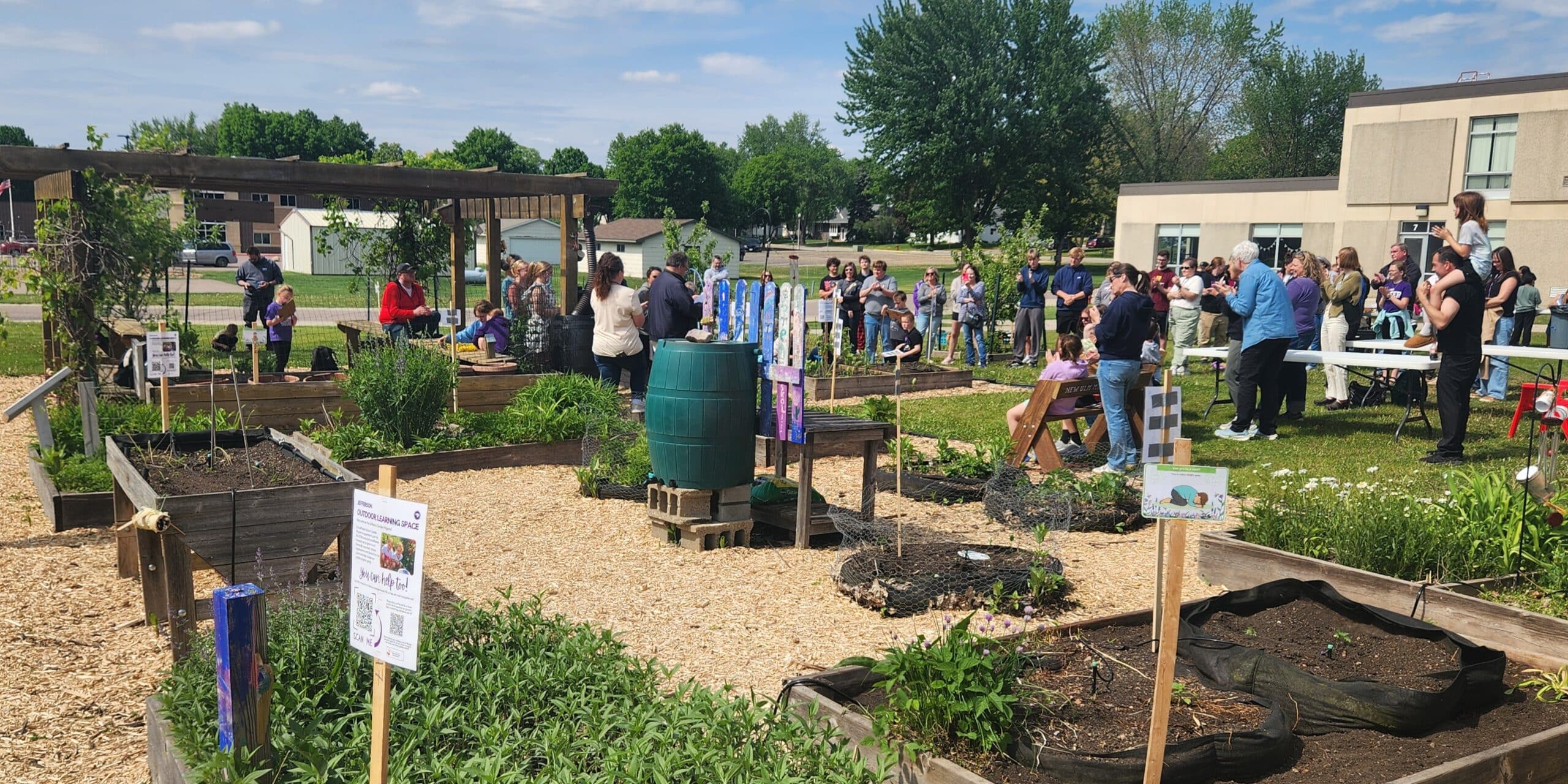 Multiple garden beds with various green plants surrounding a center area with wood chips and a rain barrel. A group of people gathered in the background to listen to someone speak.