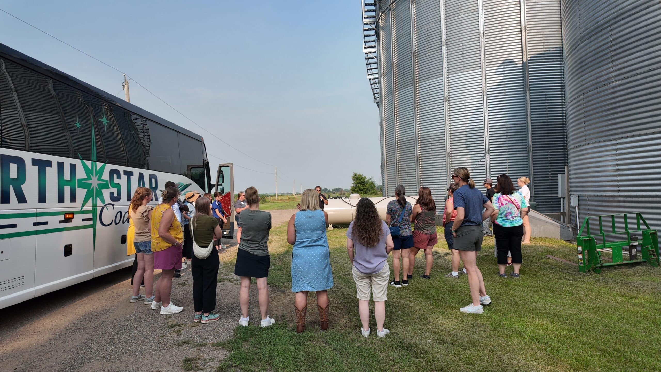 Group of people standing in a semi-circle between a coach bus and silos, listening to a man with a microphone