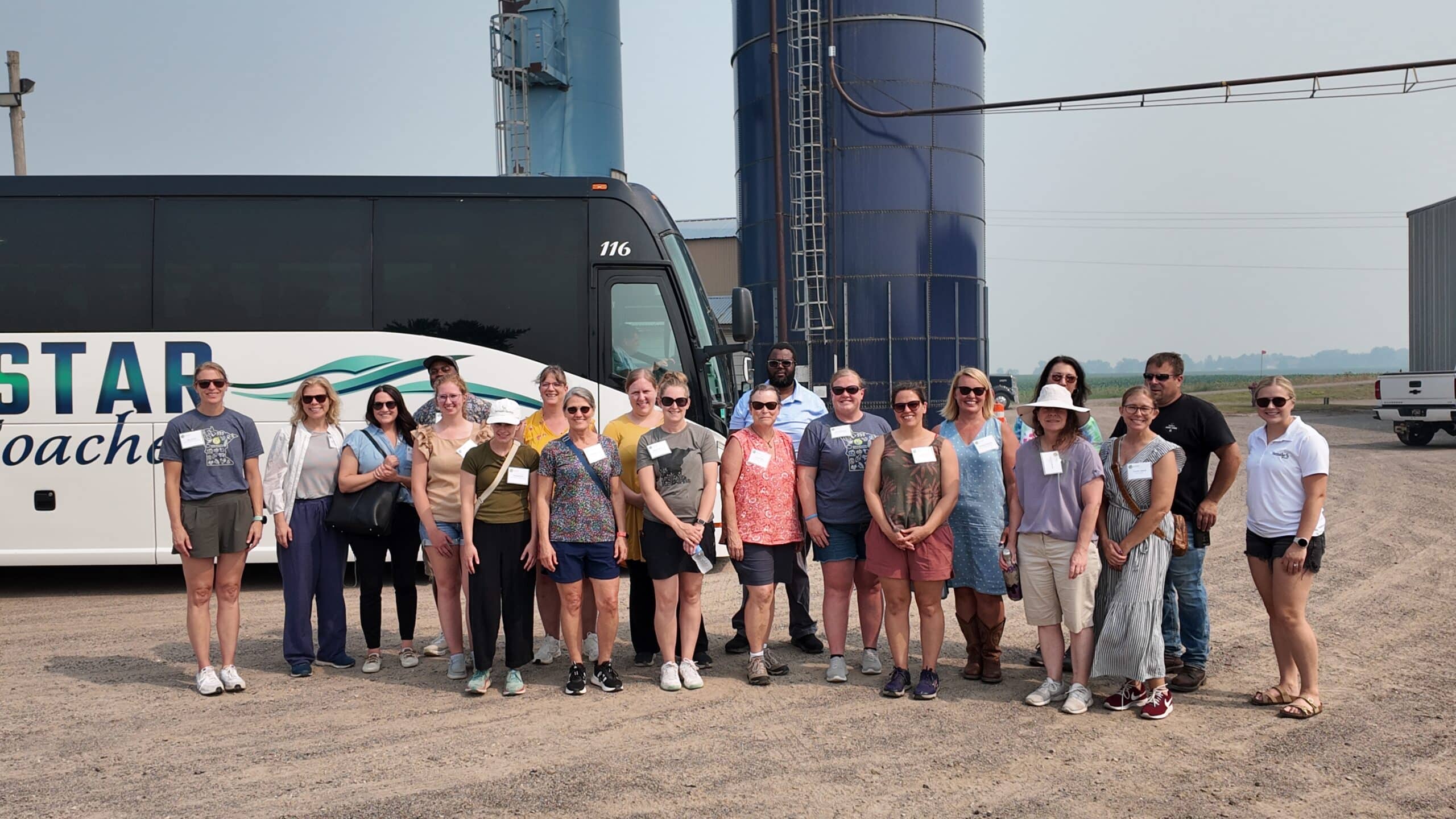 A group of about 20 people posing for a photo in front of a coach bus, a silo stands tall in the background