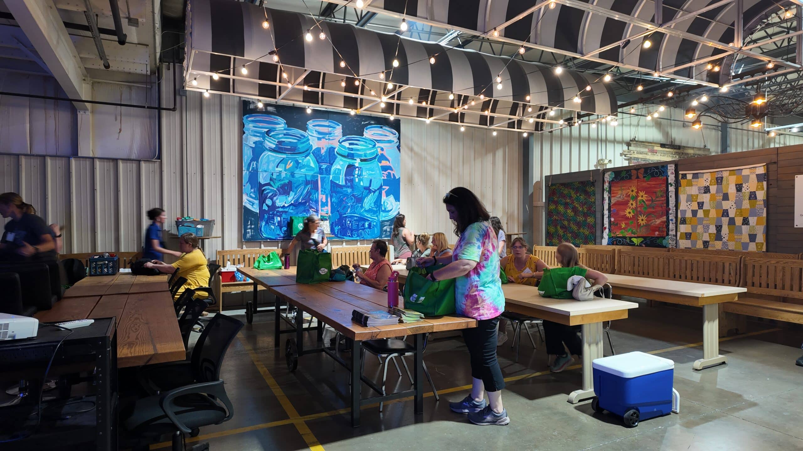 In a warehouse space, educators gather at wooden tables, string lights hang overhead, a large blue painting of mason jars hangs on the wall