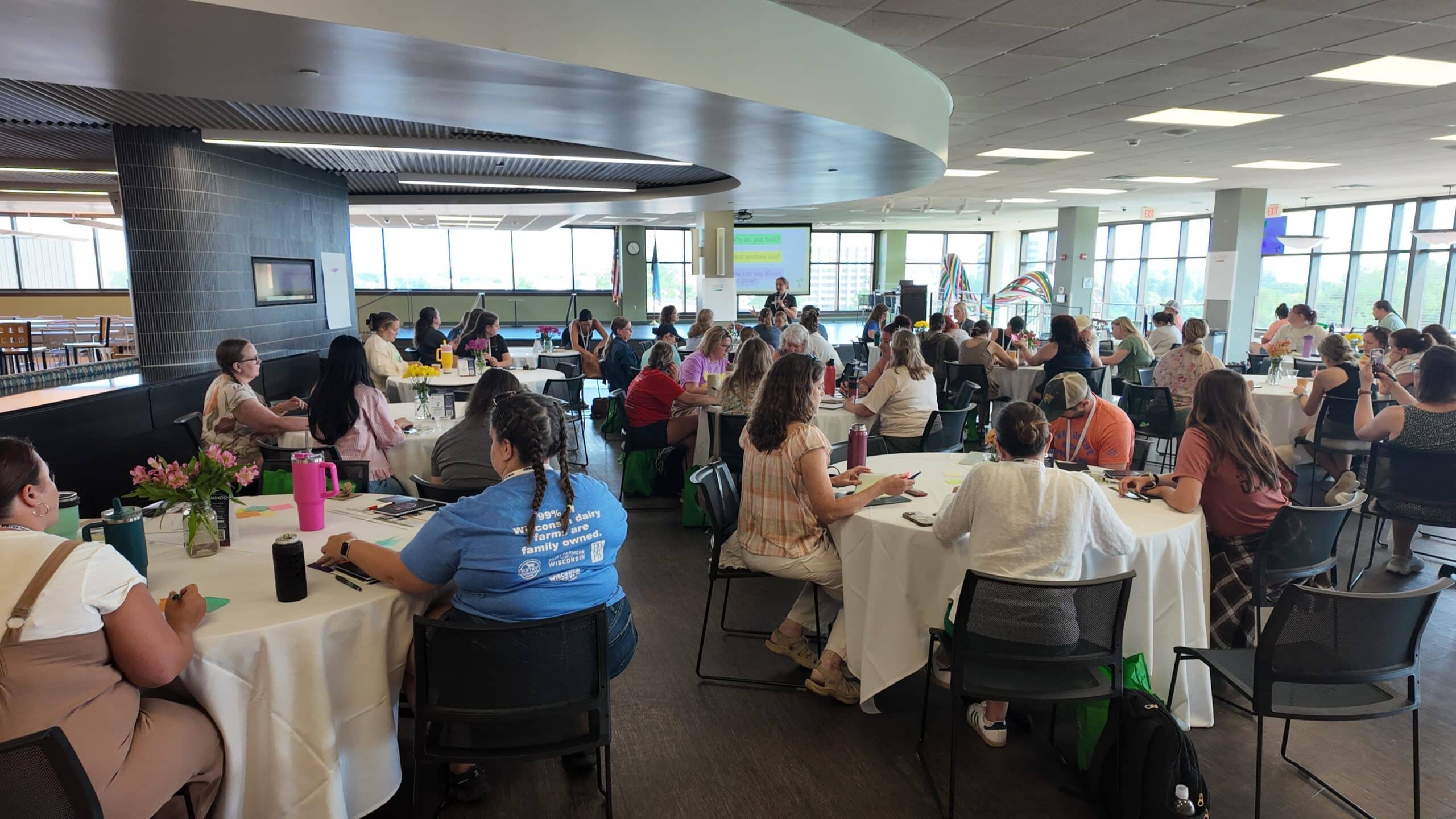 A group of people fill a room sitting at round tables, writing on sticky notes