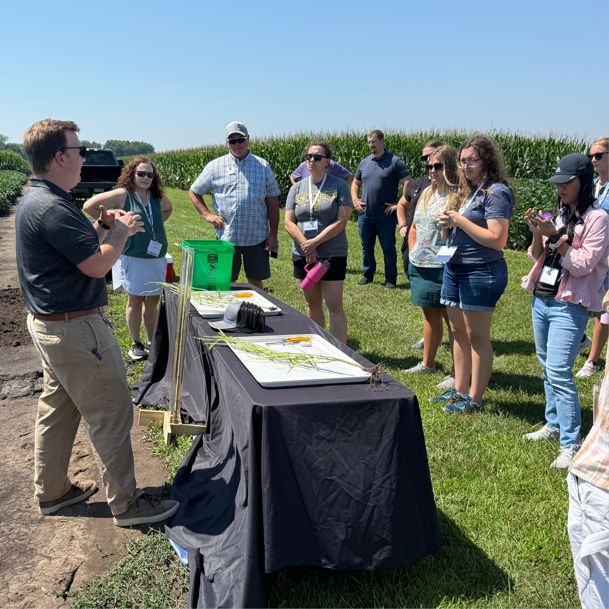 Man behind a table with trays of corn plant sprouts speaking to a small group outside in a field next to corn