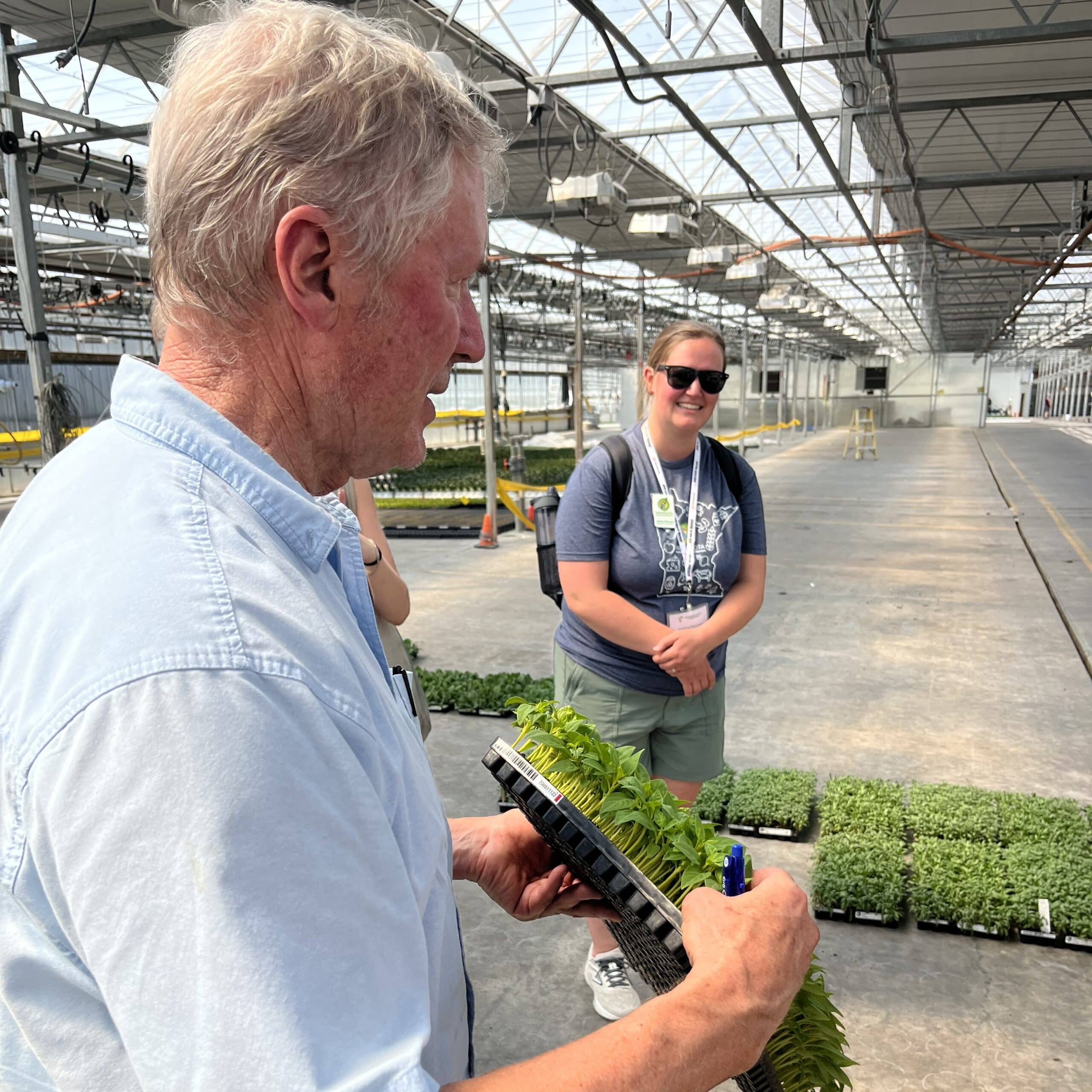Older man holding a tray of small plants while an onlooker listens