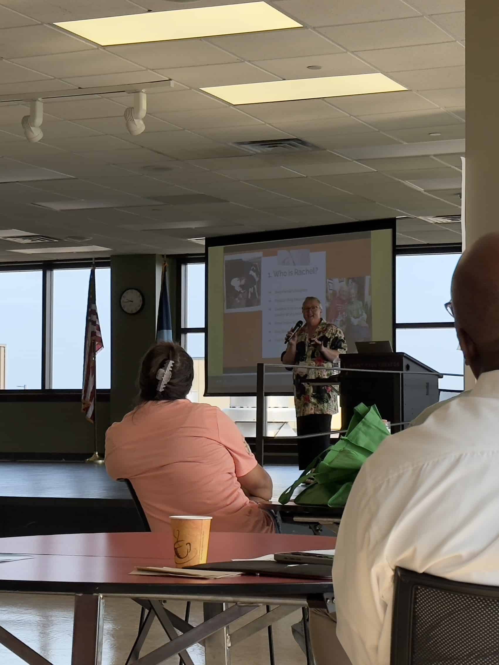 A woman stands on a stage with a microphone, presenting from a slide deck, two people sit at tables listening