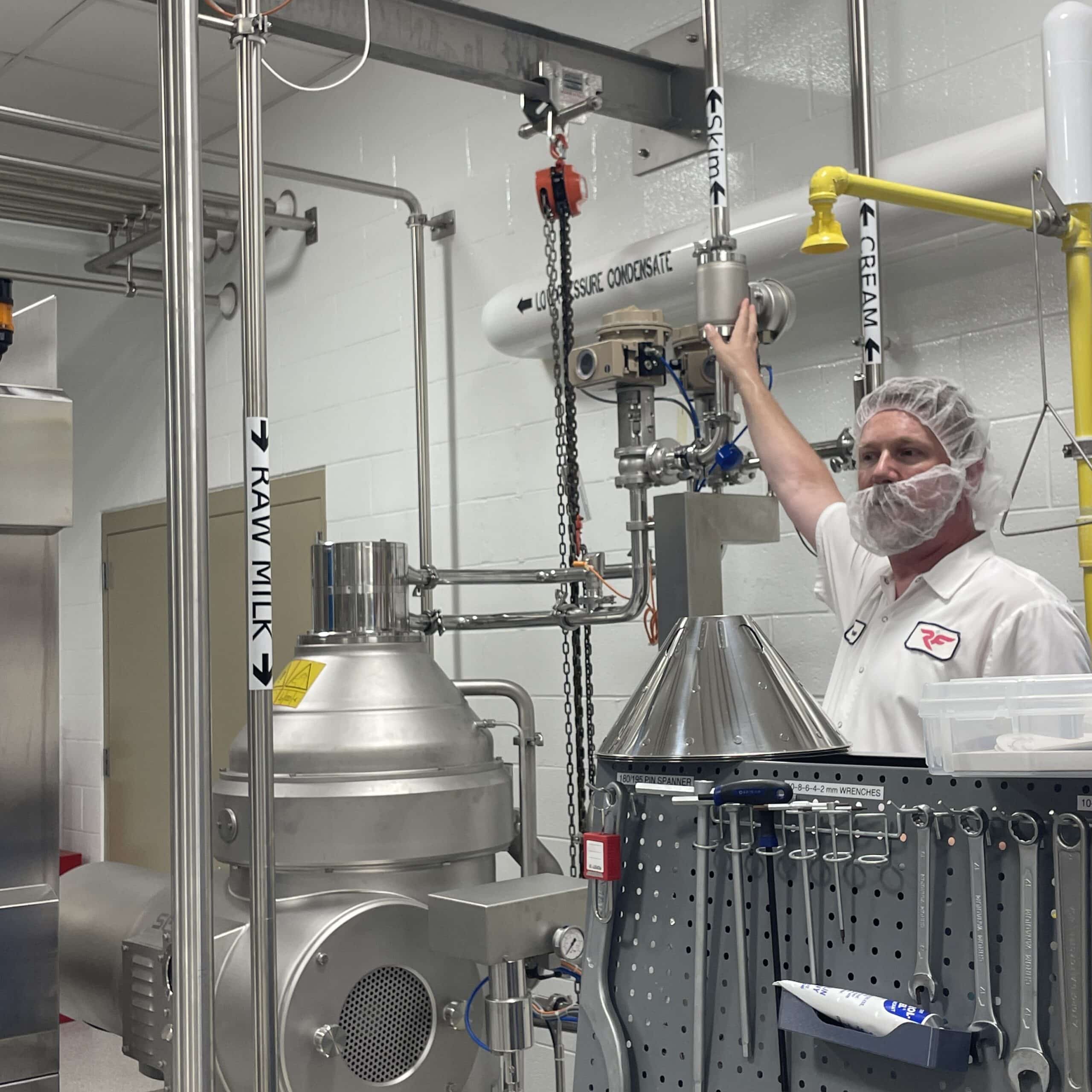 Man in white shirt and protective hair and beard nets gesturing to metal tubing labeled Skim and Cream in an industrial kitchen