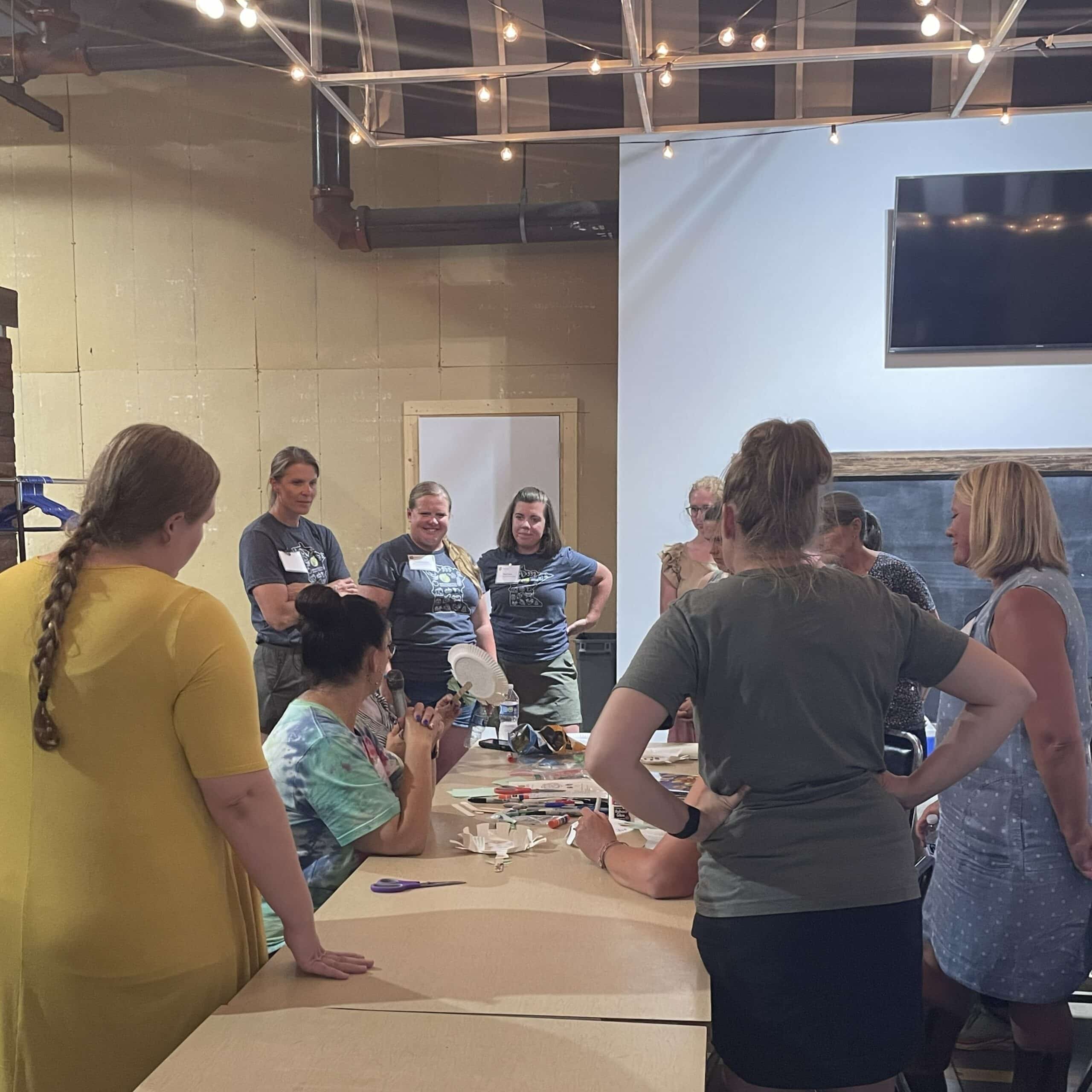 People stand around a table listening to one person talk about the lesson plan activity spread on the table