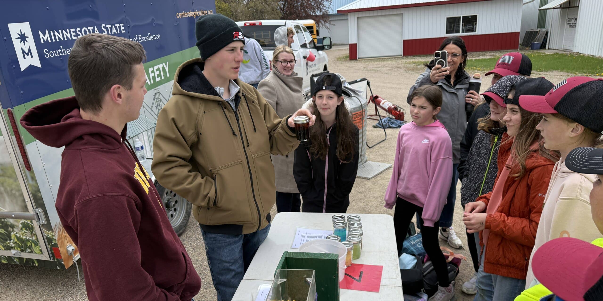 Two people stand at a white table showing a group of students some agriculture materials while outside