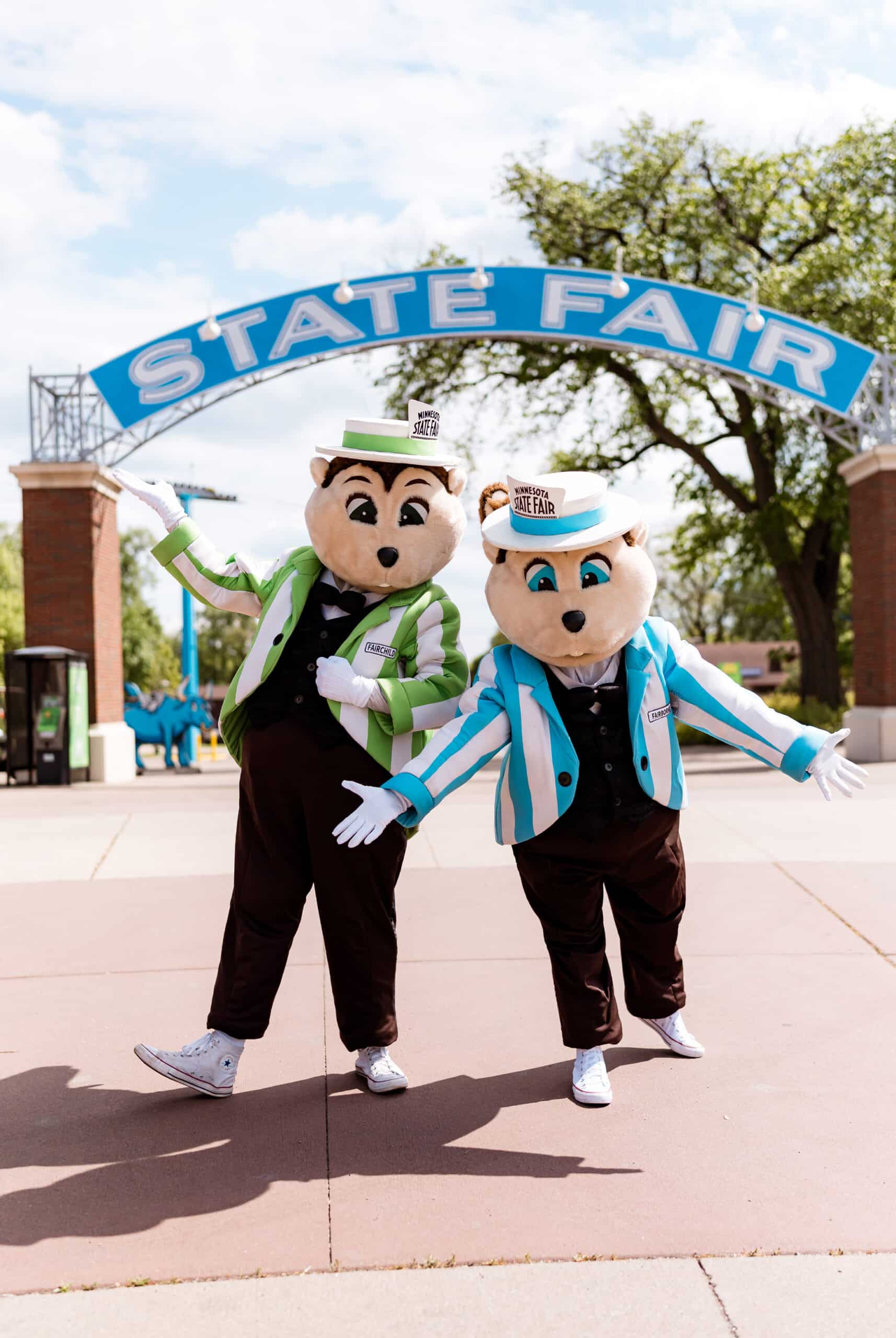 Two gopher mascots, one in a green-striped blazer and hat and one in a blue-striped blazer and hat, standing under a blue arch with the words state fair